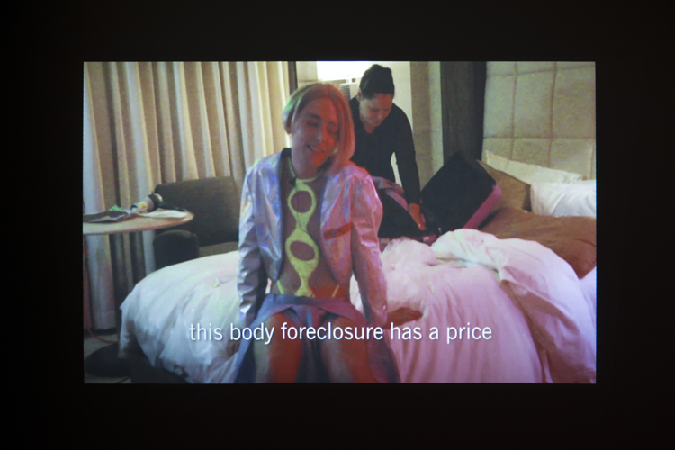 Isaac Pool A Alternatives. A video still of a white person in a shiny, skimpy pop star outfit sitting on a bed in a hotel. They are closing their eyes and smiling while a brown woman in the back unpacks a suitcase on the bed. A subtitle on the screen reads this body foreclosure has a price