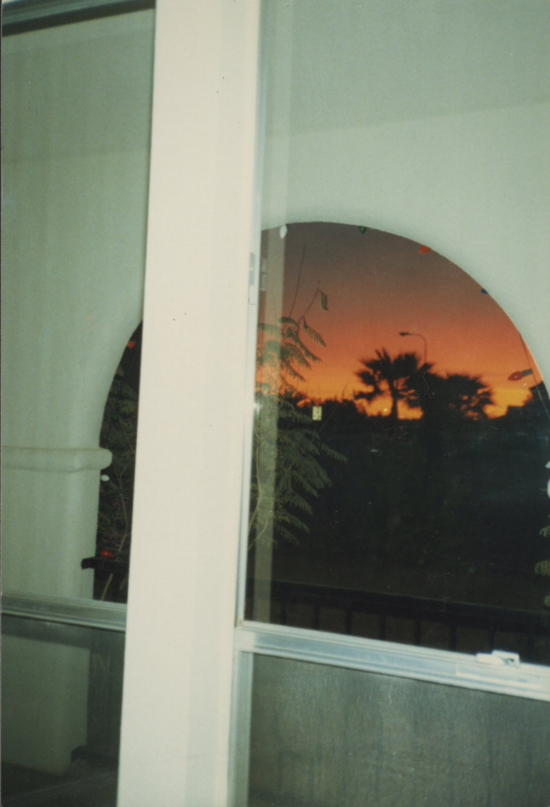 Isaac Pool, Light Stain. A photograph of a sunset over the desert taken from inside a home with the frame of a window and a porch blocking the image of the sunset.