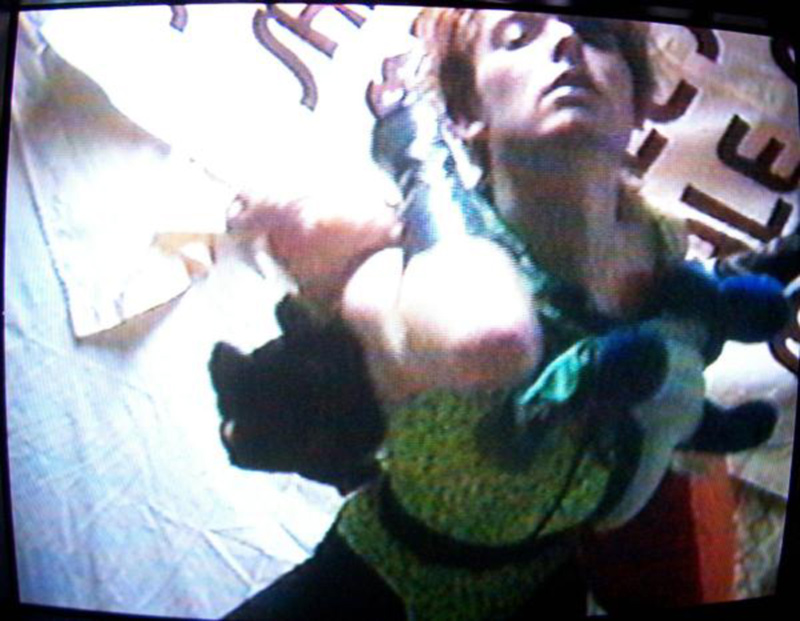 RENTAL, a still from a VHS video. A white person in a dramatic dance pose points their elbow at the camera and looks downward while wearing a long black glove and a costume made of a colorful bathing suit and stuffed animals. There is a banner in teh background that reads SALE