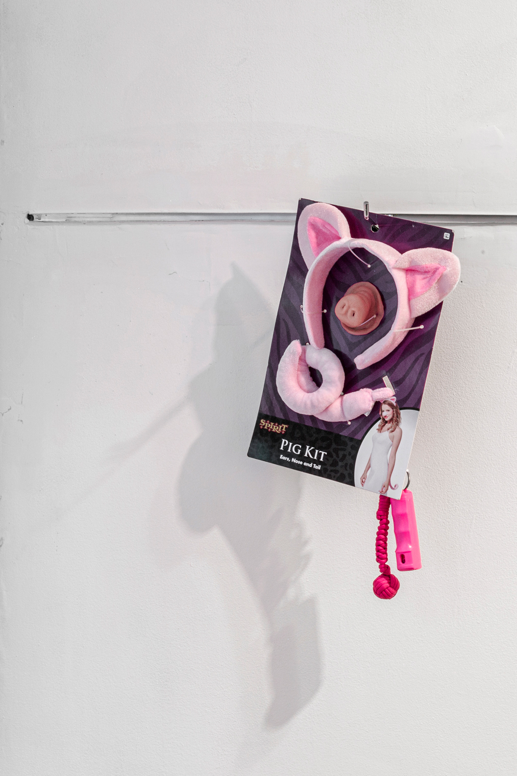 Isaac Pool A costume hanging from a retail clothing rack that's been plastered into the wall. The product is reads Pig Kit, ears, nose, and tail and all items are pink. A photograph of a white woman wearing a white dress standing in a sexy side pose wearing the costume is printed on the corner. Two pink-colored self-defense tools, a weighted monkey ball and small pepper spray gun, dangle from the corner.